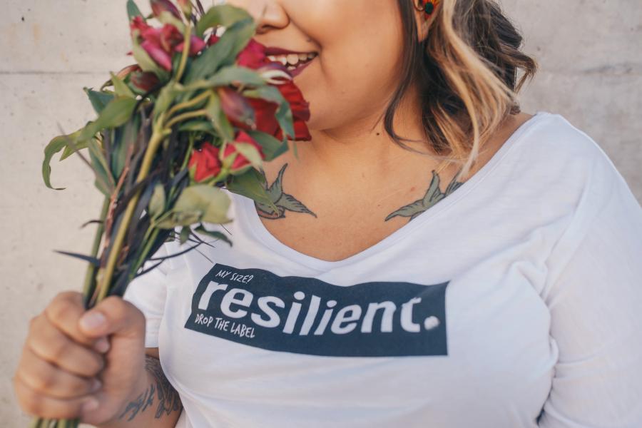 Woman smiling, holding flowers, and wearing a t-shirt that reads "My size? Resilient. Drop the label."